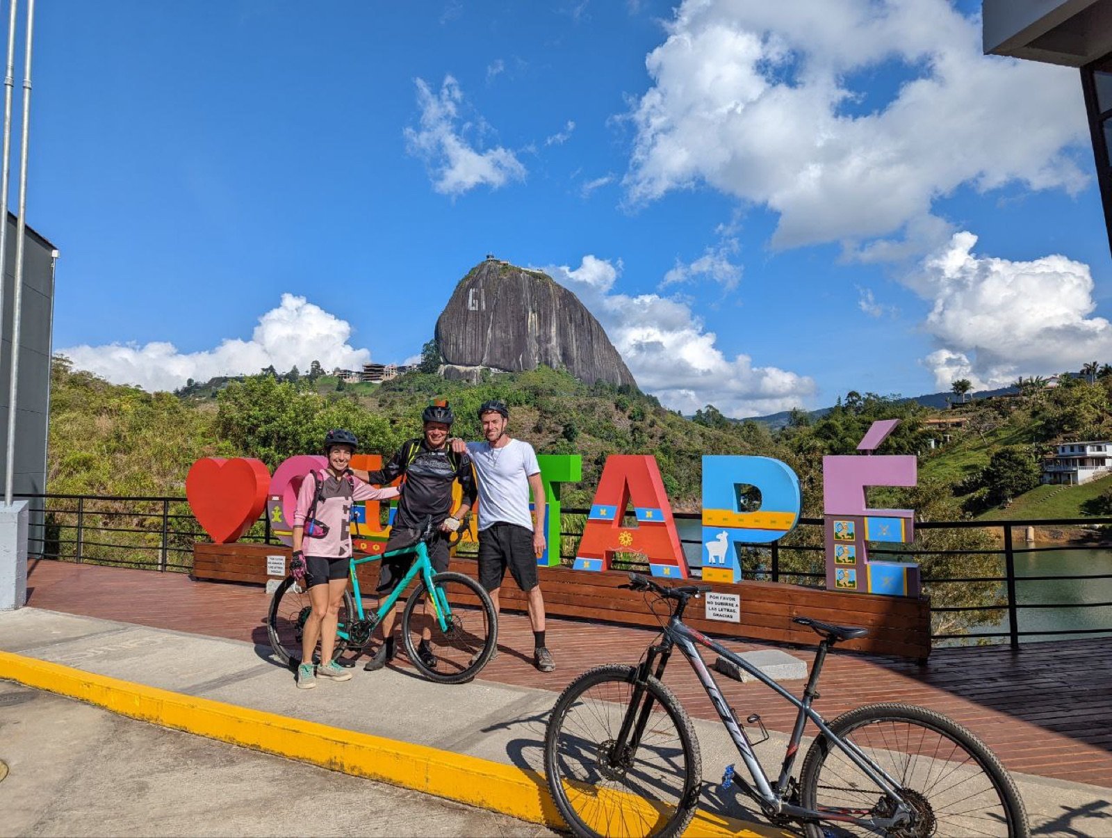 Riders with bikes beside the Guatapé town sign on the waterfront with La Piedra del Peñol behind