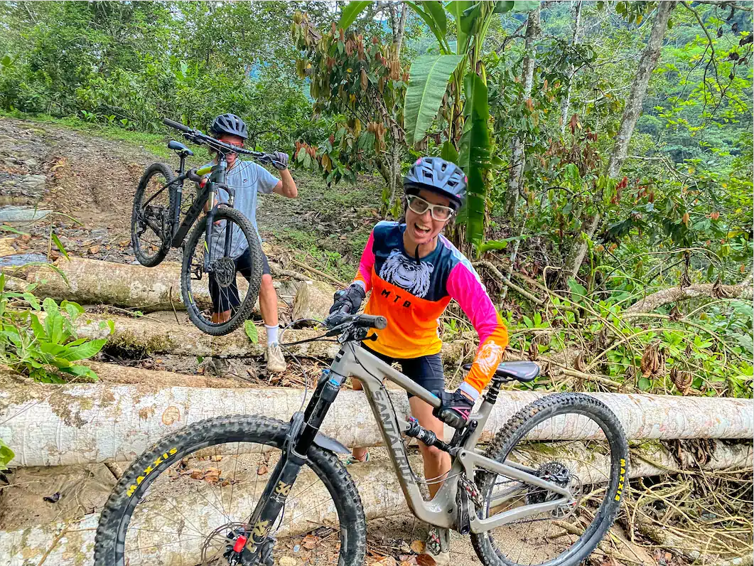 Smiling mountain biker beside a fallen-log obstacle on a lush forest trail, with another rider carrying a bike behind